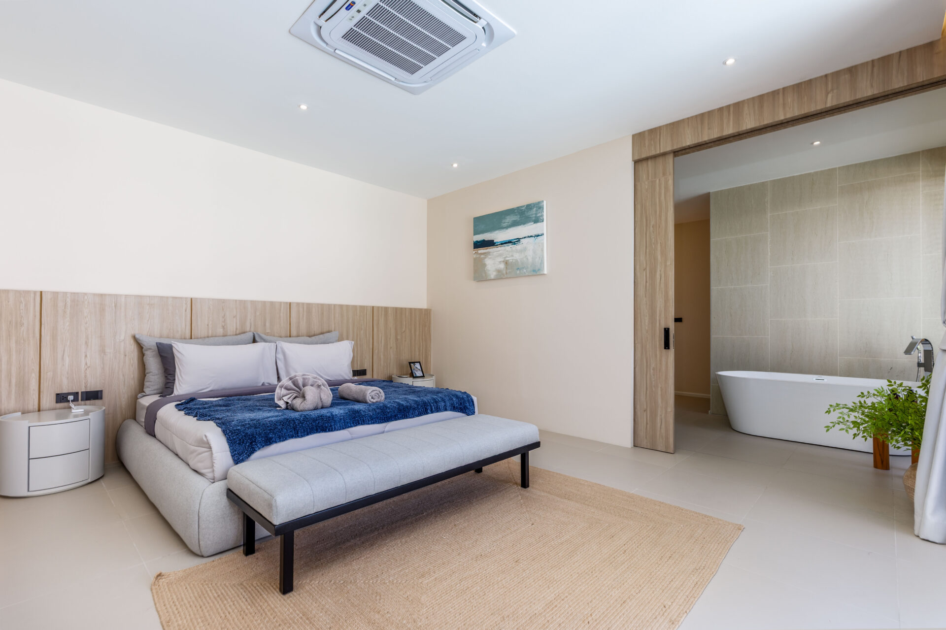 Modern hotel bedroom with a king bed, beige headboard, blue throw, and an open doorway to a tiled bathroom with a freestanding tub.