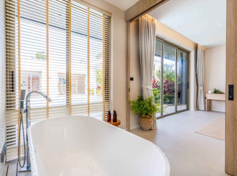 Bright bathroom with a white freestanding tub, large windows with horizontal blinds, and sliding glass doors opening to a garden.