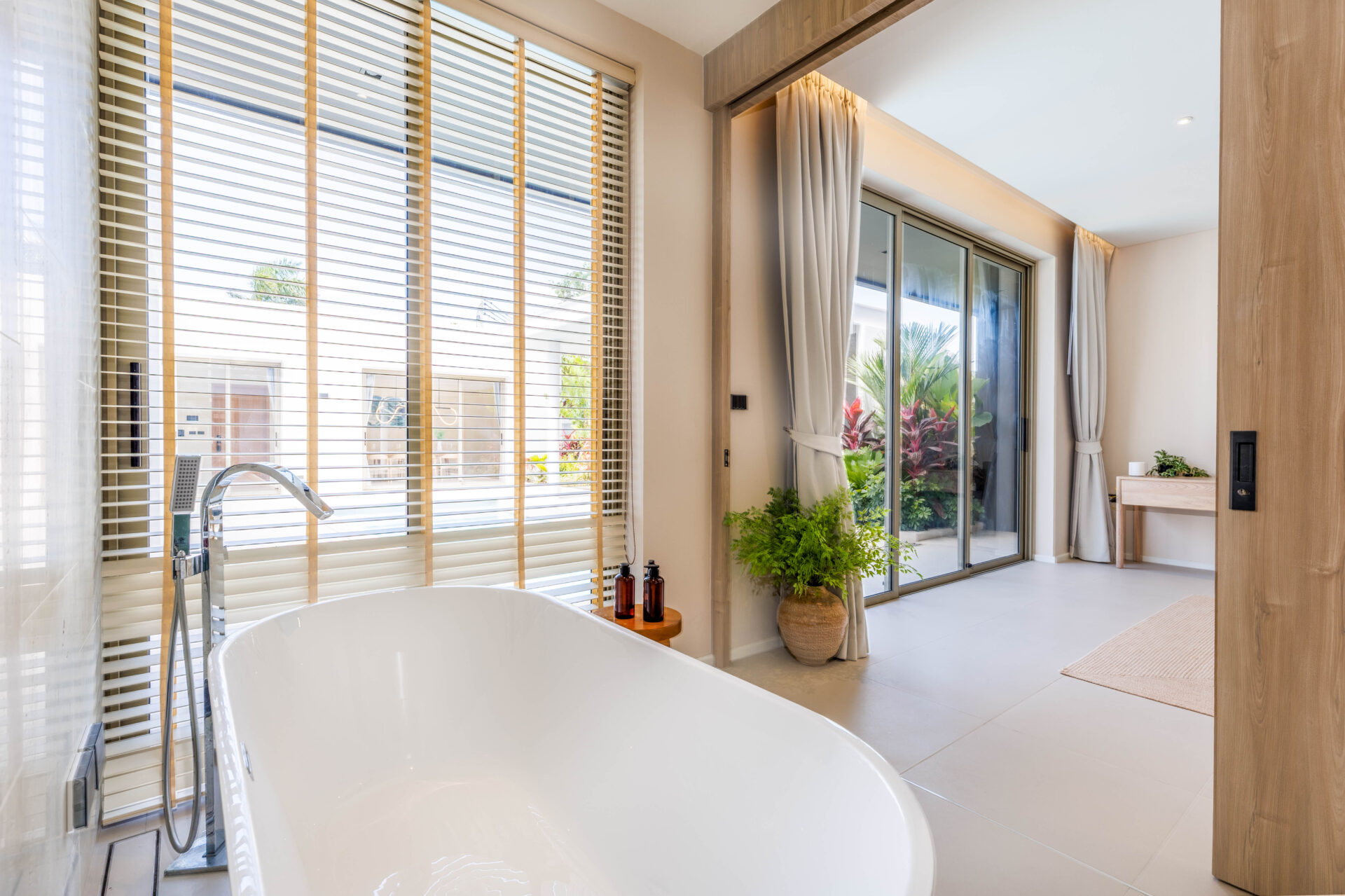 Bright bathroom with a white freestanding tub, large windows with horizontal blinds, and sliding glass doors opening to a garden.