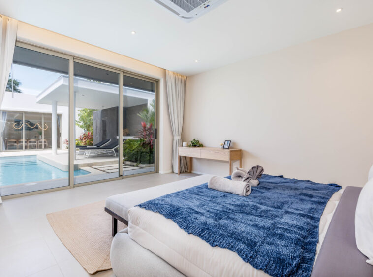 Bright modern bedroom with sliding glass doors to a pool patio, bed with a blue throw and neatly rolled towels at the foot of the bed.