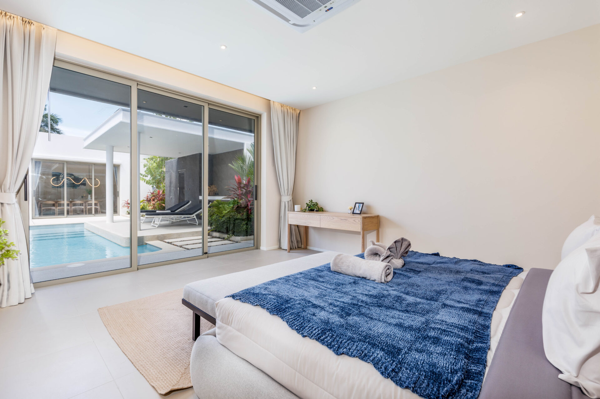 Bright modern bedroom with sliding glass doors to a pool patio, bed with a blue throw and neatly rolled towels at the foot of the bed.