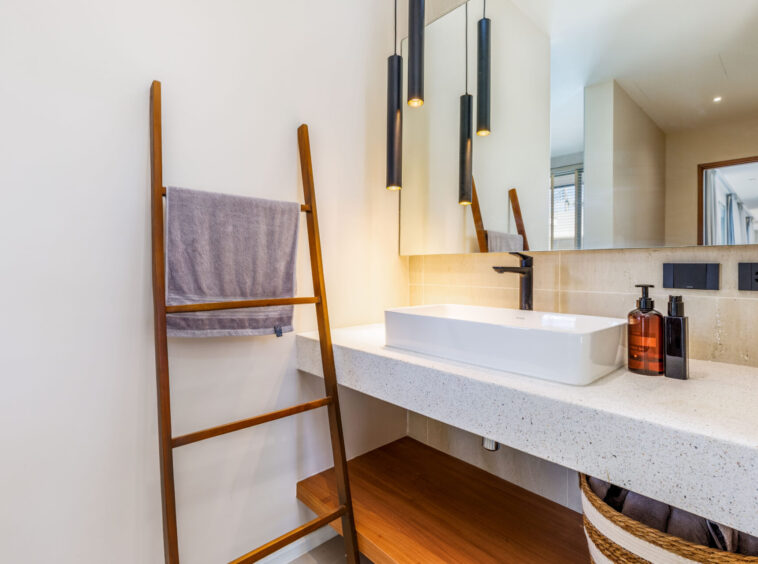 Modern bathroom with a white rectangular sink, black faucet, beige counter, pendant lights, a wooden towel ladder, and a woven basket.