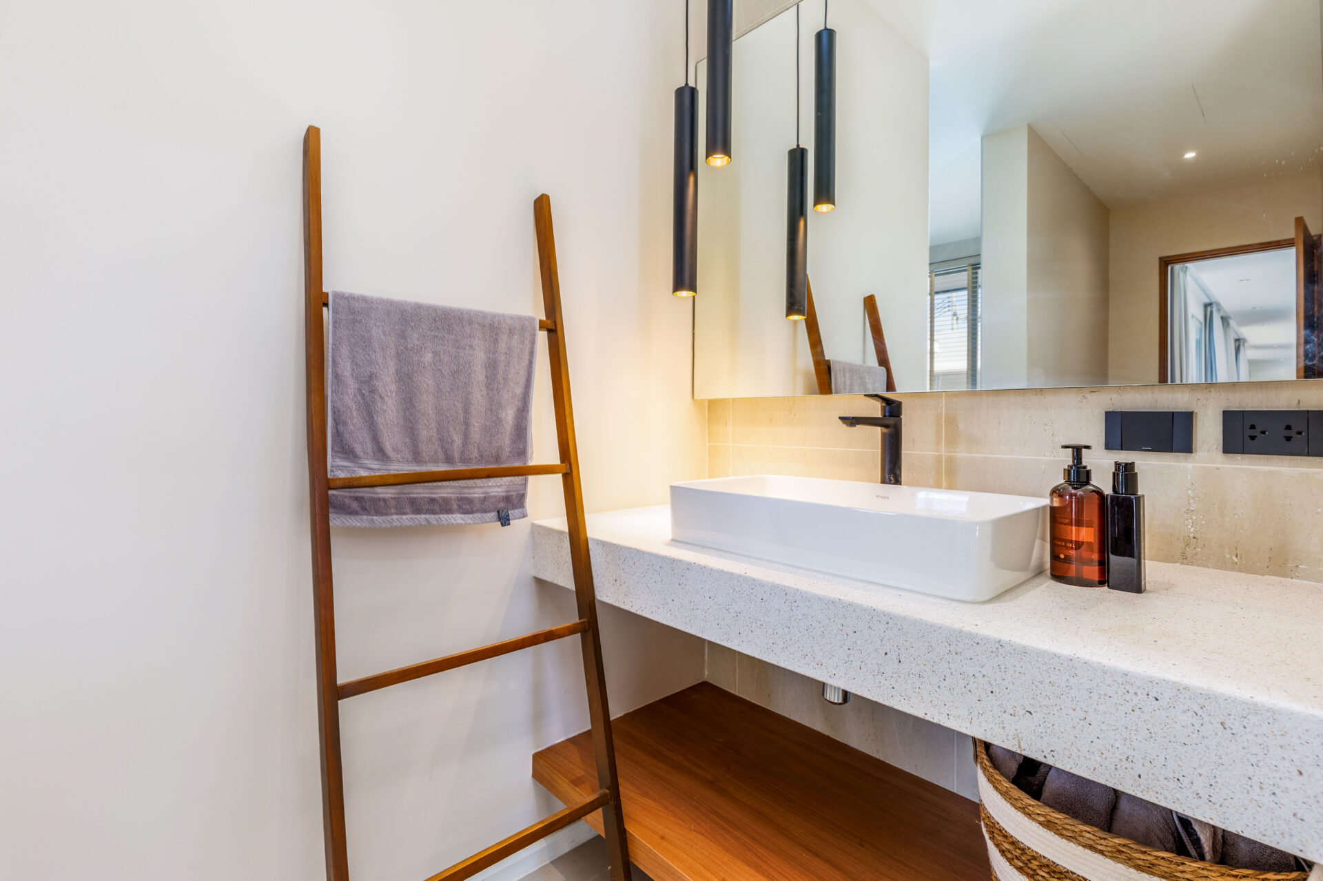 Modern bathroom with a white rectangular sink, black faucet, beige counter, pendant lights, a wooden towel ladder, and a woven basket.