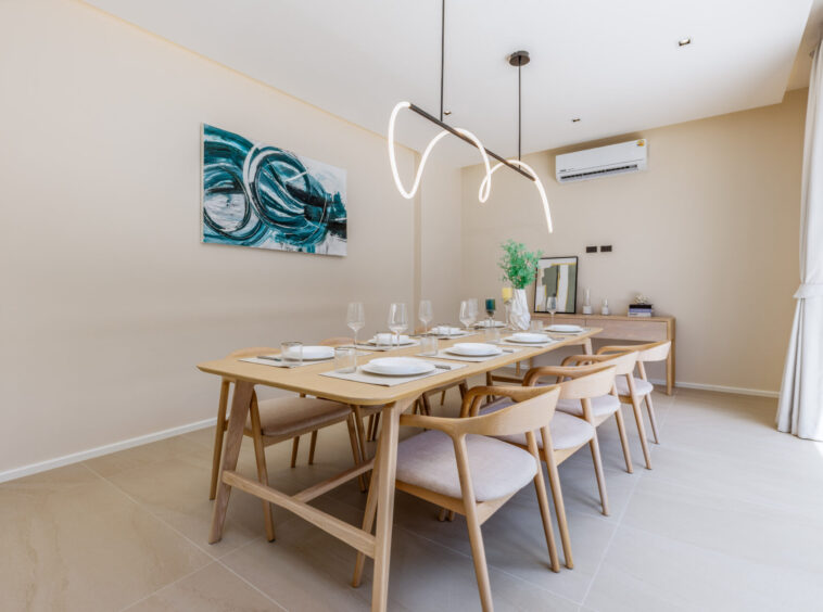 Modern dining room with a long wooden table set for eight, a sculptural pendant light, and an abstract teal artwork on a beige wall | the space feels bright and contemporary, ready for a meal served with glassware and plates to set the scene.