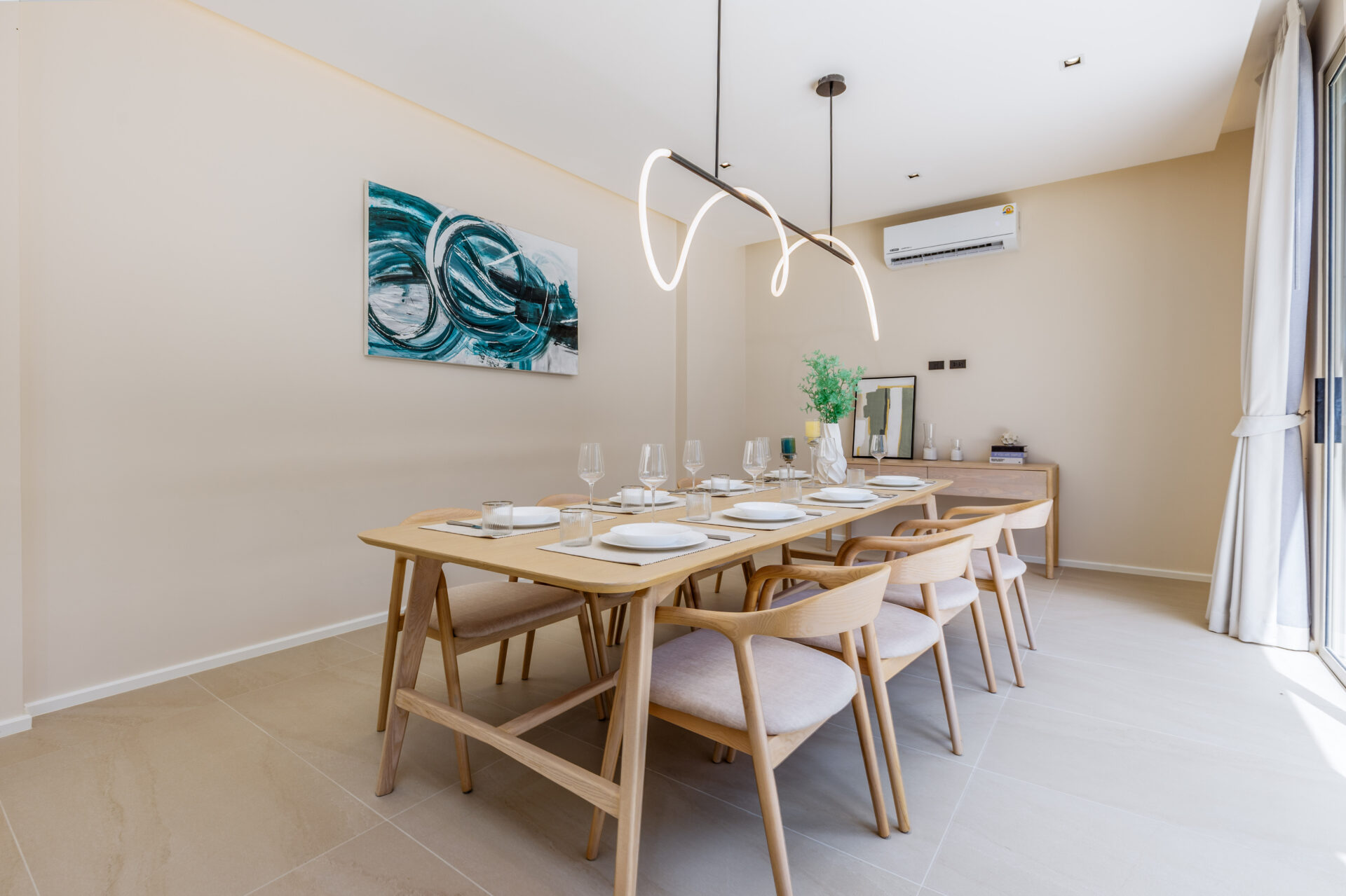 Modern dining room with a long wooden table set for eight, a sculptural pendant light, and an abstract teal artwork on a beige wall | the space feels bright and contemporary, ready for a meal served with glassware and plates to set the scene.