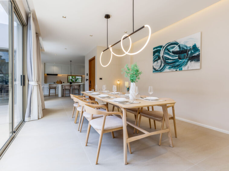 Long communal dining table set for a meal in a bright, modern kitchen-dining area with circular pendant lights and a teal abstract painting on the wall