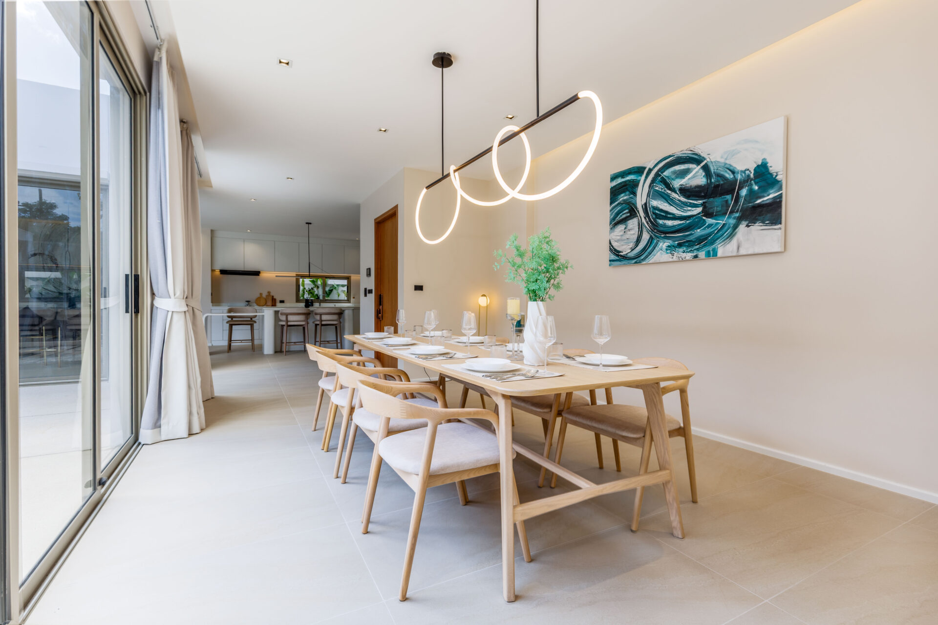 Long communal dining table set for a meal in a bright, modern kitchen-dining area with circular pendant lights and a teal abstract painting on the wall