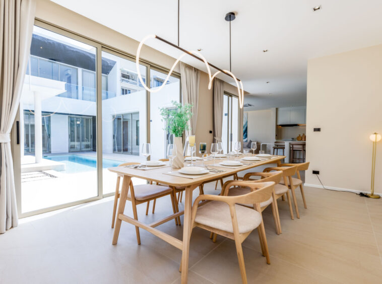 Bright dining room with a long wooden table set for a meal, beige chairs, and a modern looping pendant light; floor-to-ceiling windows overlook a pool courtyard.