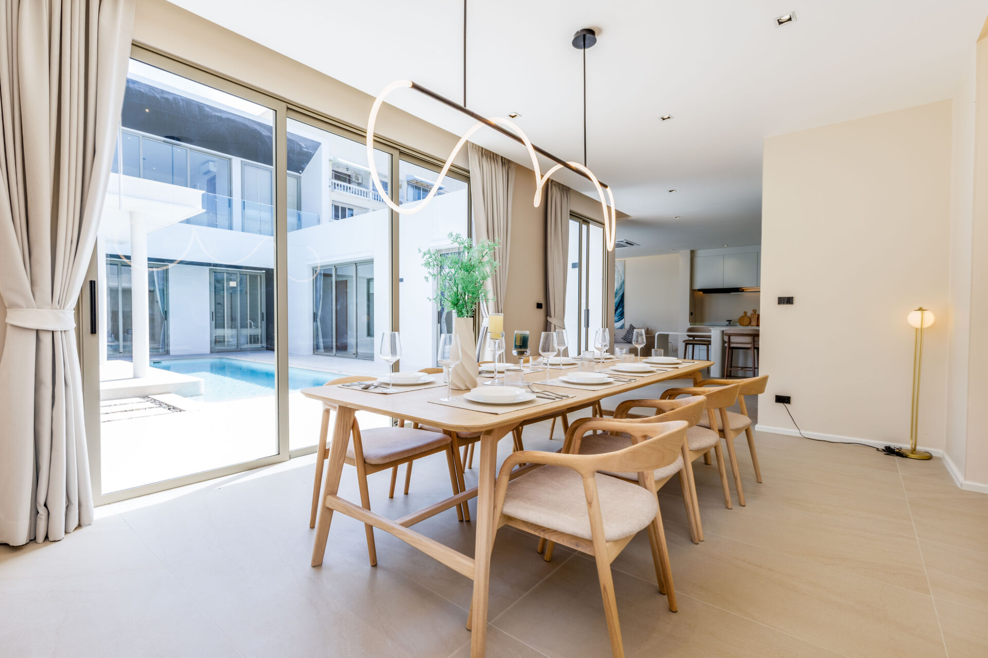 Bright dining room with a long wooden table set for a meal, beige chairs, and a modern looping pendant light; floor-to-ceiling windows overlook a pool courtyard.