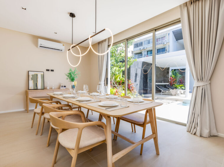 Bright dining room with a long light wood table set for six, by large glass doors and a circular ring chandelier above.