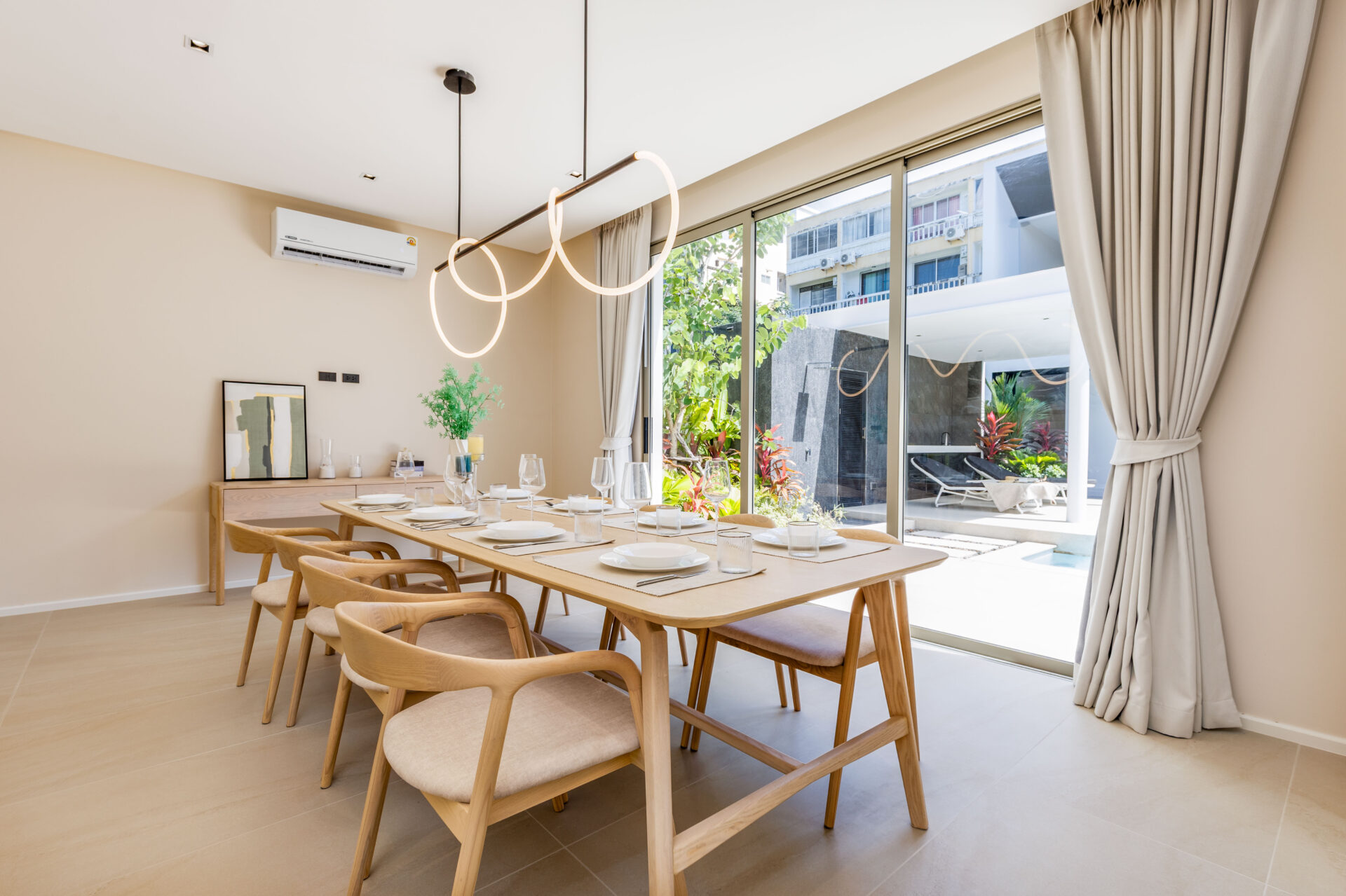 Bright dining room with a long light wood table set for six, by large glass doors and a circular ring chandelier above.
