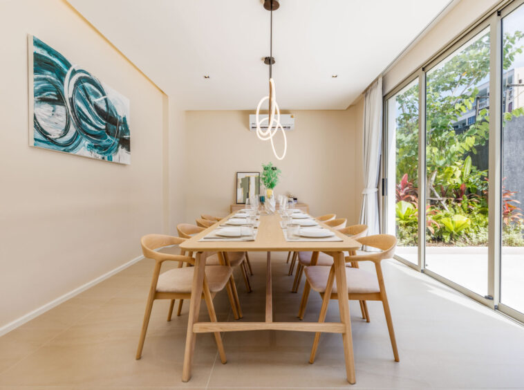 Long wooden dining table set for a meal in a bright dining room with large glass doors opening to a lush garden outside.
