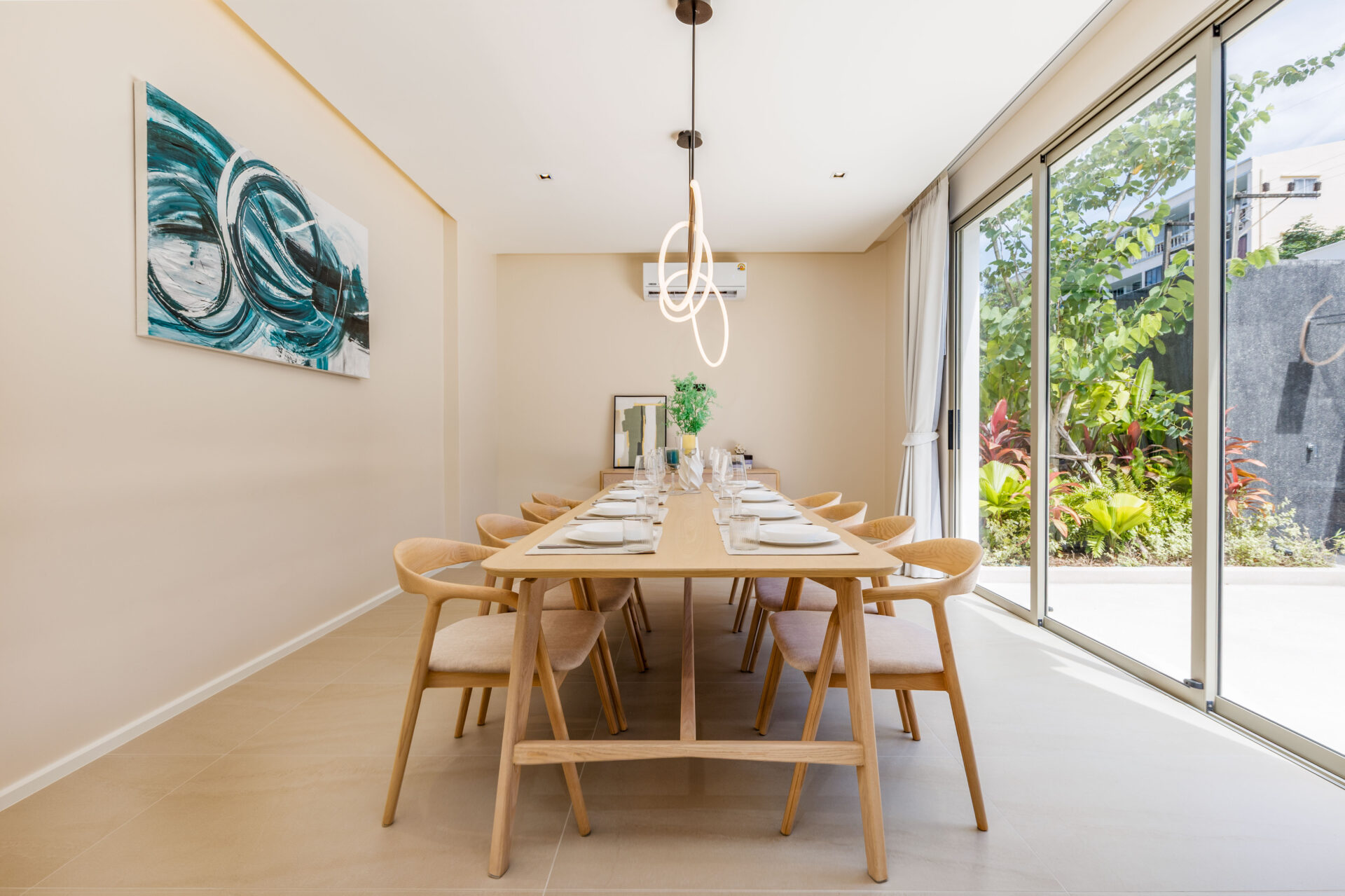Long wooden dining table set for a meal in a bright dining room with large glass doors opening to a lush garden outside.