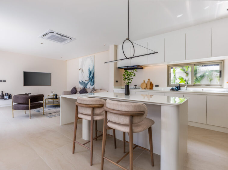 Open-plan kitchen and living area with white cabinets, a marble light-toned island, beige bar stools, and a wall-mounted TV in the living room.