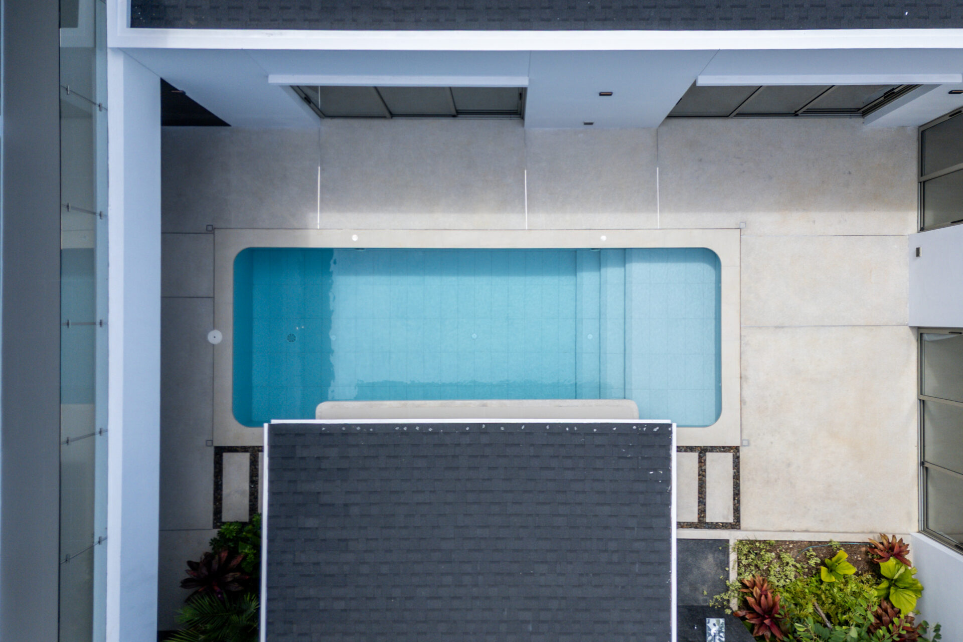 Aerial view of a rectangular rooftop pool surrounded by light-colored walls and dark shaded panels.