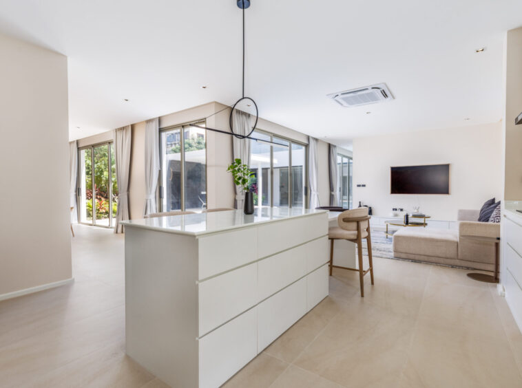 Bright open-plan living area with a white kitchen island, beige floors, and large glass doors to the outdoors.