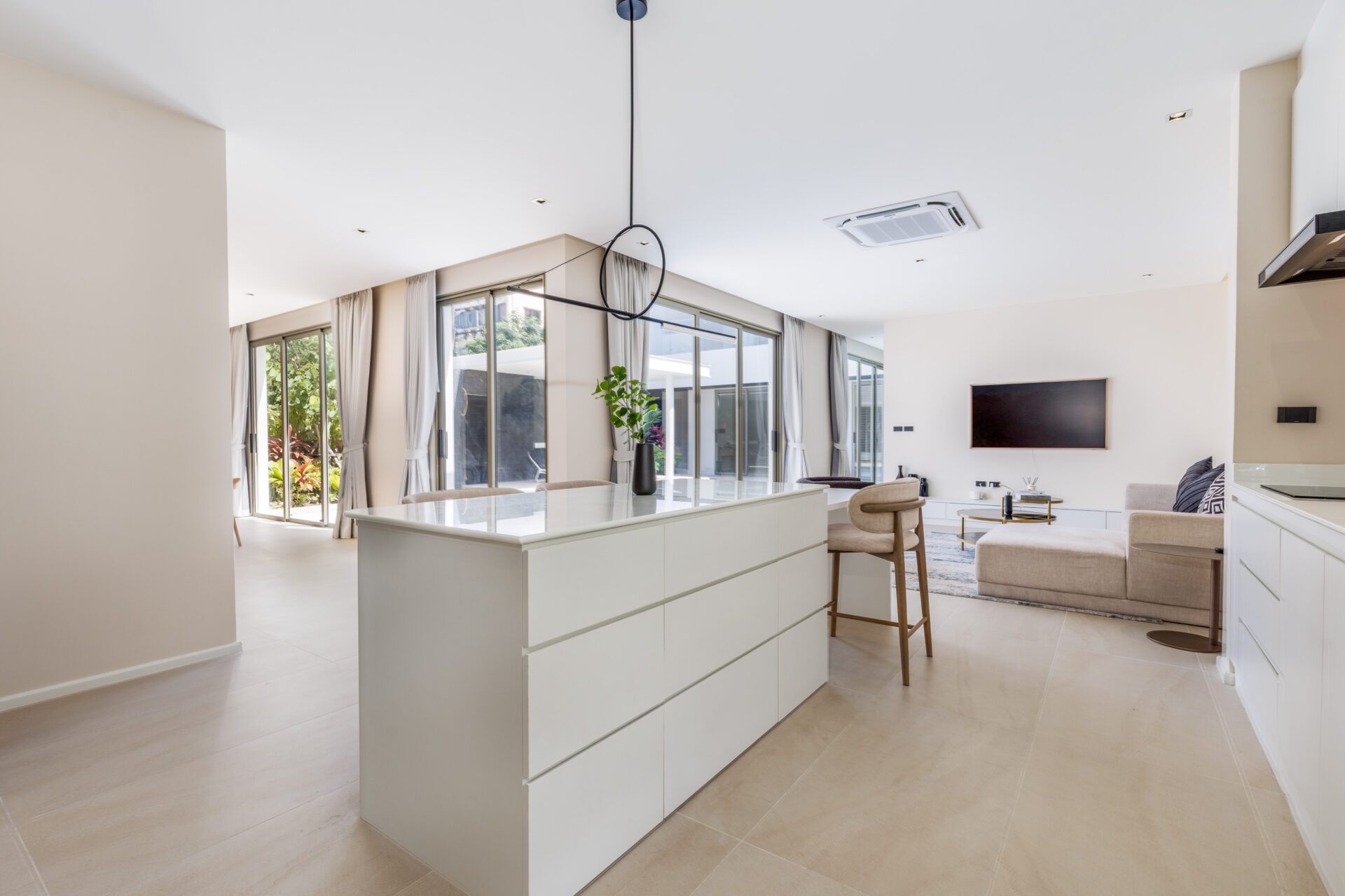 Bright open-plan living area with a white kitchen island, beige floors, and large glass doors to the outdoors.