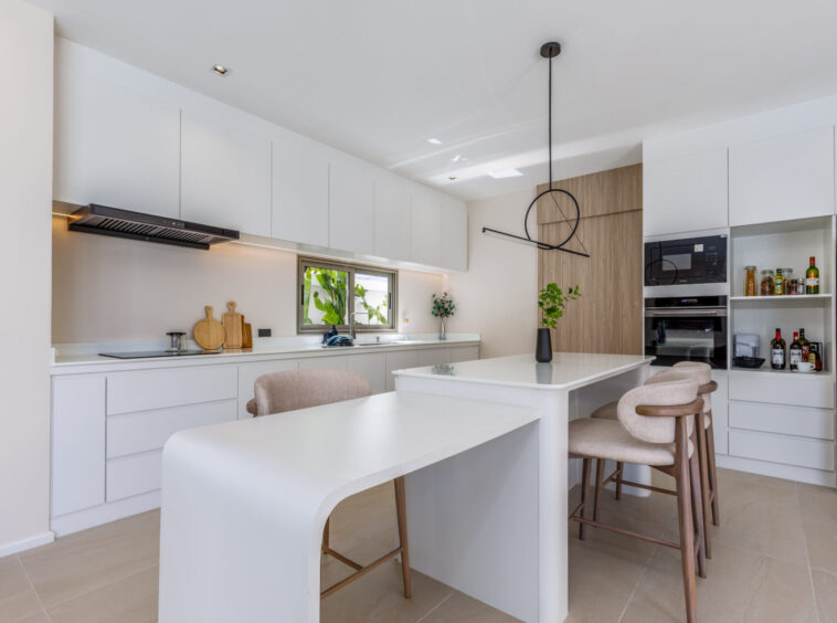 Bright white modern kitchen with a large island, beige bar stools, and a wall of cabinets and built-in oven.