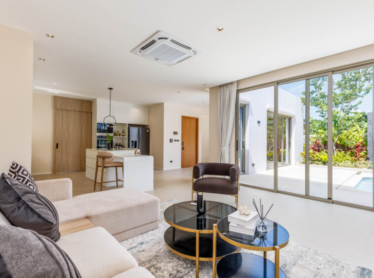 Bright, open-concept living area with beige sectional sofa, round black-and-gold coffee tables, and a view of the outdoor courtyard through large sliding glass doors. A modern kitchen island and pantry are in the background.