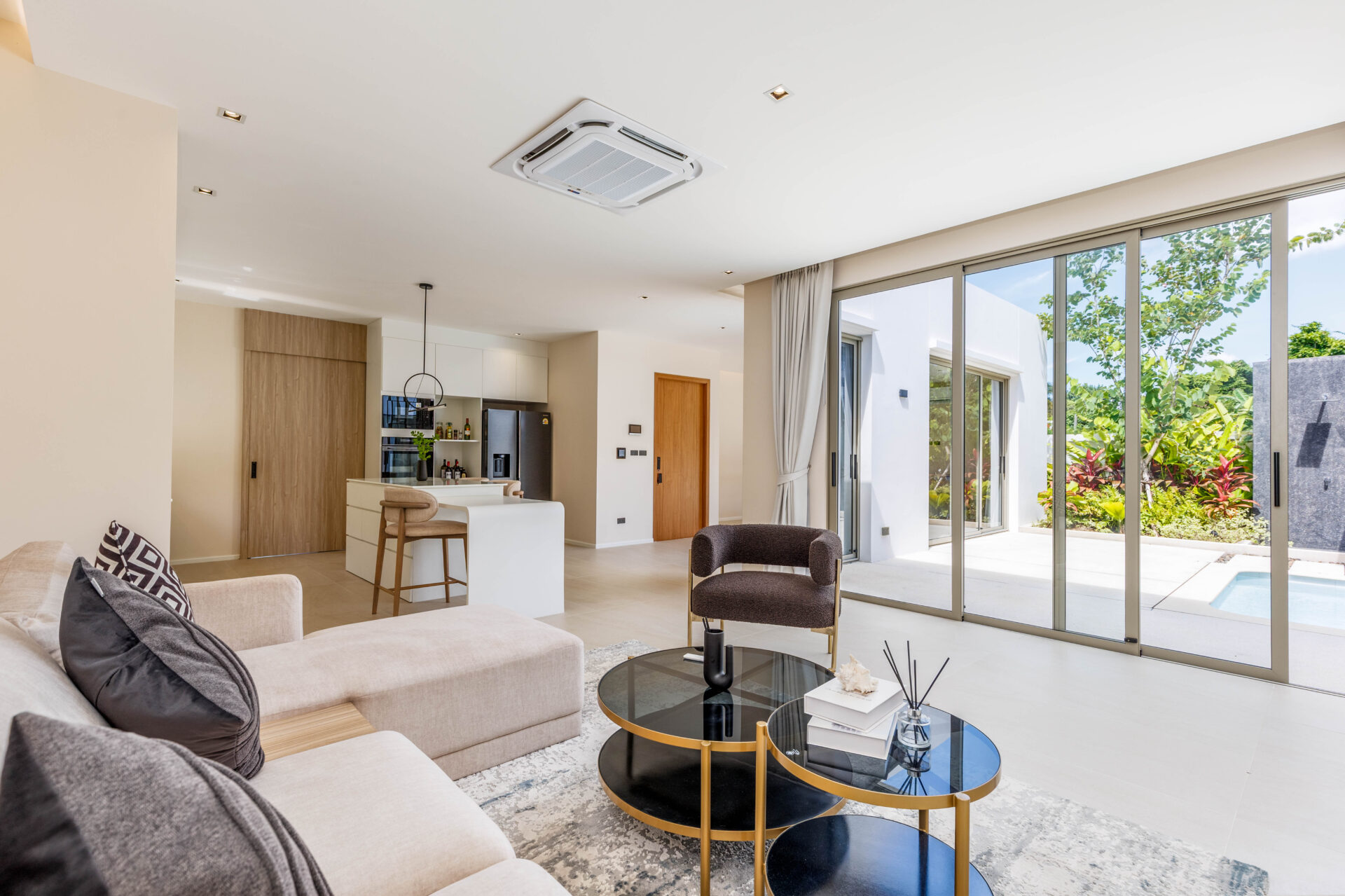 Bright, open-concept living area with beige sectional sofa, round black-and-gold coffee tables, and a view of the outdoor courtyard through large sliding glass doors. A modern kitchen island and pantry are in the background.