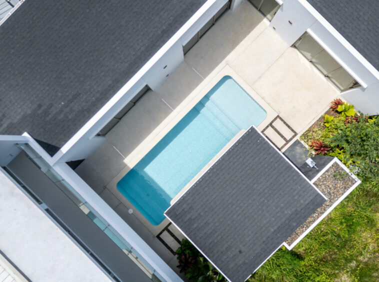 Aerial view of a modern house complex with a rectangular turquoise pool surrounded by white walls and dark gray roof tiles, and greenery on the side.