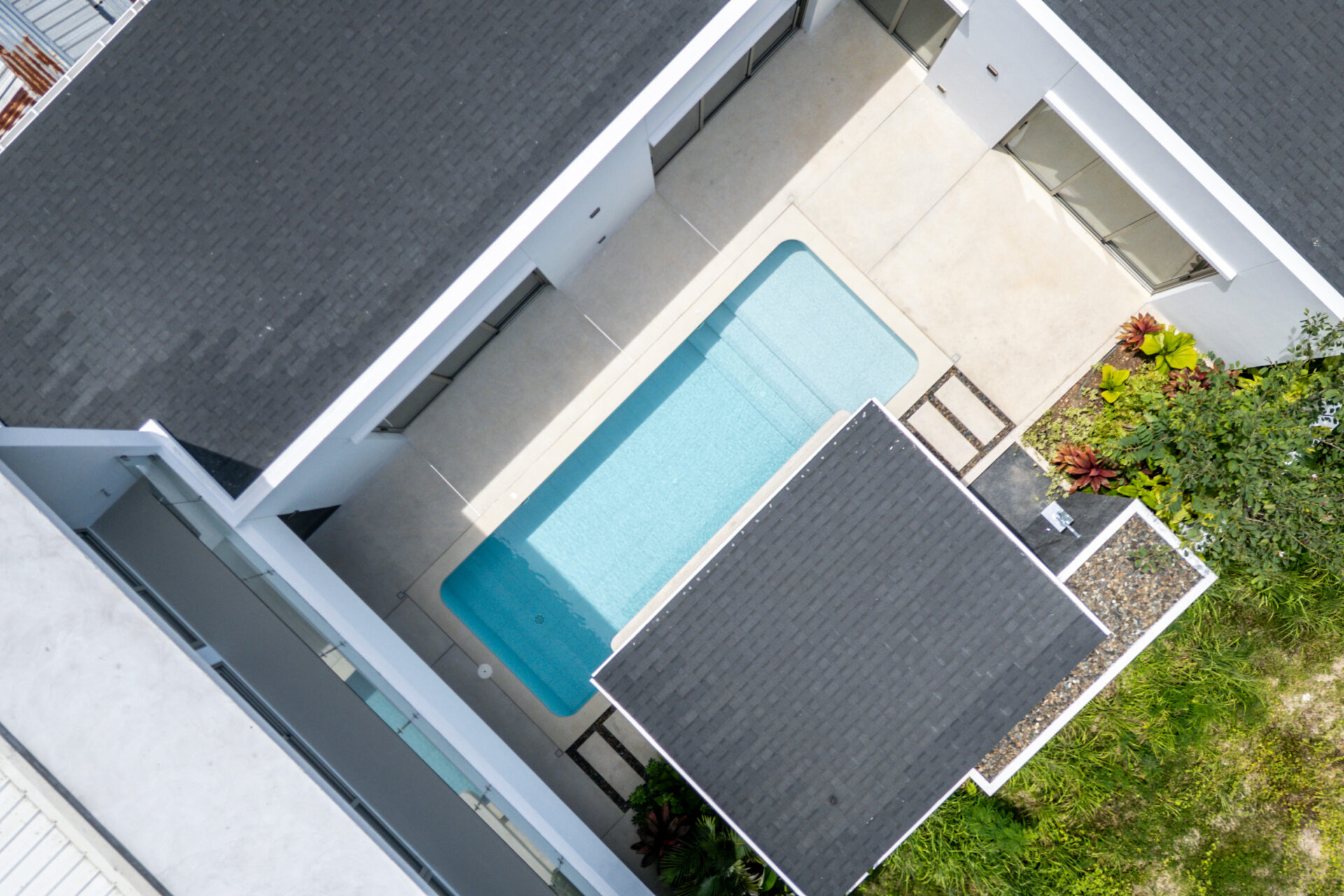 Aerial view of a modern house complex with a rectangular turquoise pool surrounded by white walls and dark gray roof tiles, and greenery on the side.