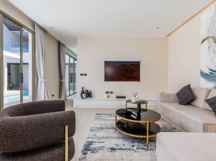 Bright modern living room with large windows overlooking a pool, a beige sectional sofa, and dark brown textured chairs.