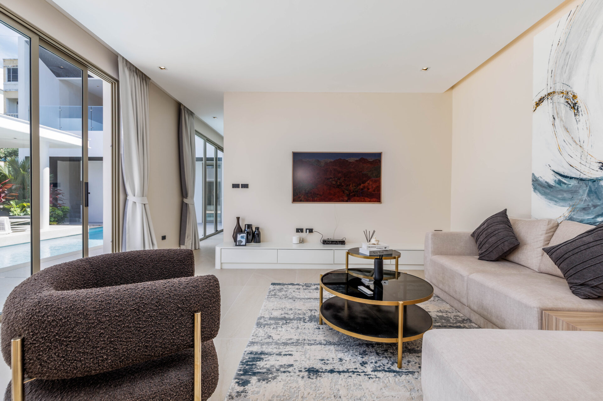 Bright modern living room with large windows overlooking a pool, a beige sectional sofa, and dark brown textured chairs.