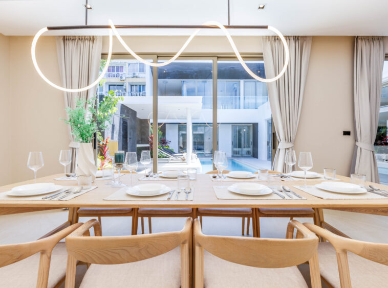 Long wooden dining table set for a formal meal with white plates, glassware and beige chairs, by large windows overlooking a courtyard and pool.