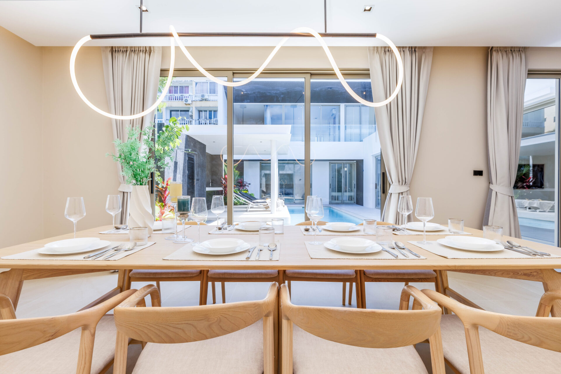 Long wooden dining table set for a formal meal with white plates, glassware and beige chairs, by large windows overlooking a courtyard and pool.