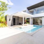 Modern house with a rectangular swimming pool, white patio, and glass balcony railing; lounge chairs under a shaded canopy.
