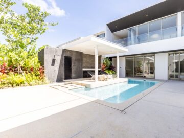 Modern house with a rectangular swimming pool, white patio, and glass balcony railing; lounge chairs under a shaded canopy.