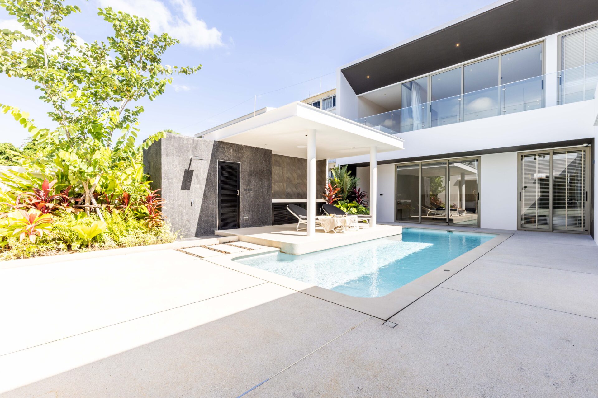 Modern house with a rectangular swimming pool, white patio, and glass balcony railing; lounge chairs under a shaded canopy.