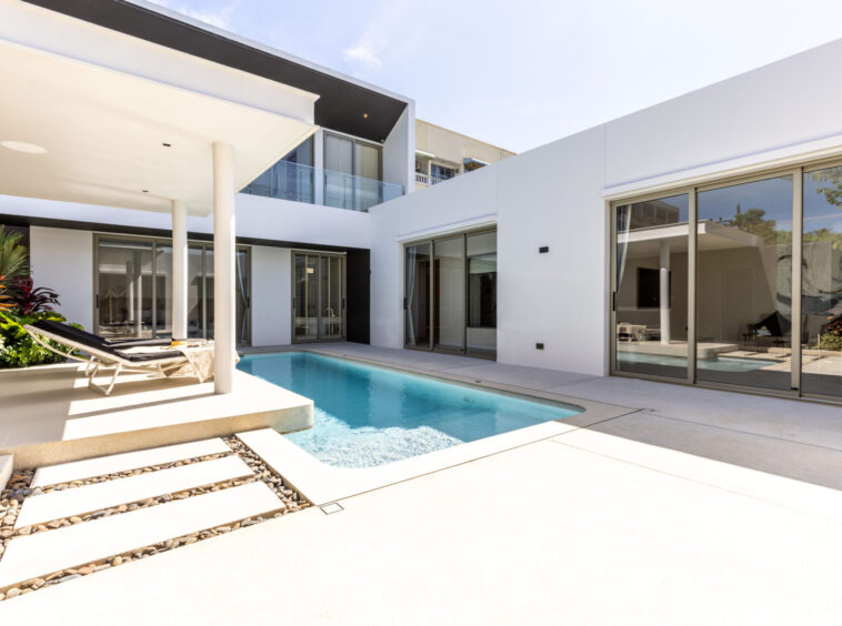 Modern white villa with glass doors surrounding a rectangular pool and a lounge chair on a sunny patio.