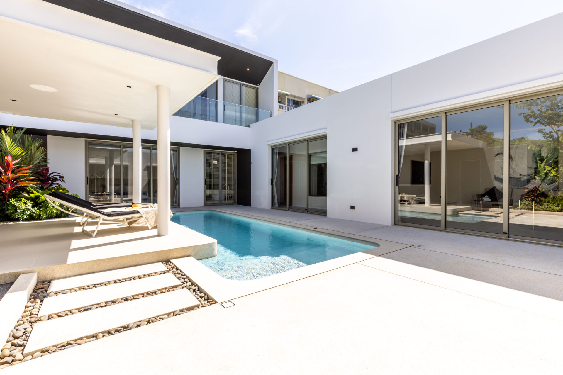 Modern white villa with glass doors surrounding a rectangular pool and a lounge chair on a sunny patio.