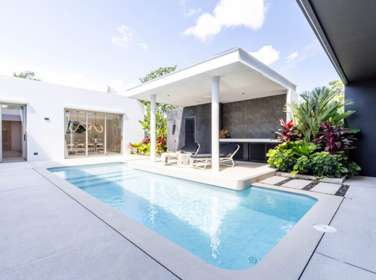 Modern backyard pool area with white patio, lounge chairs, and a shaded outdoor seating/kitchen zone under a white canopy.