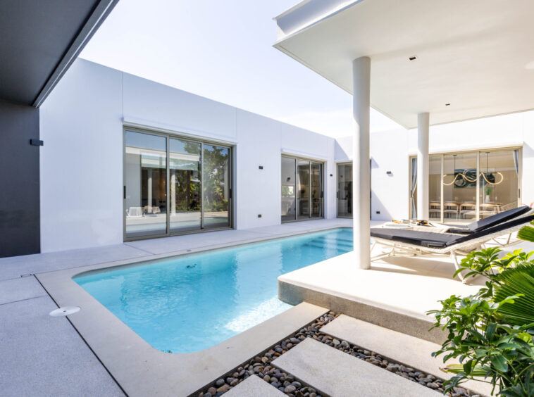 Modern white house with a rectangular pool, glass sliding doors, and sun loungers along a covered patio area.