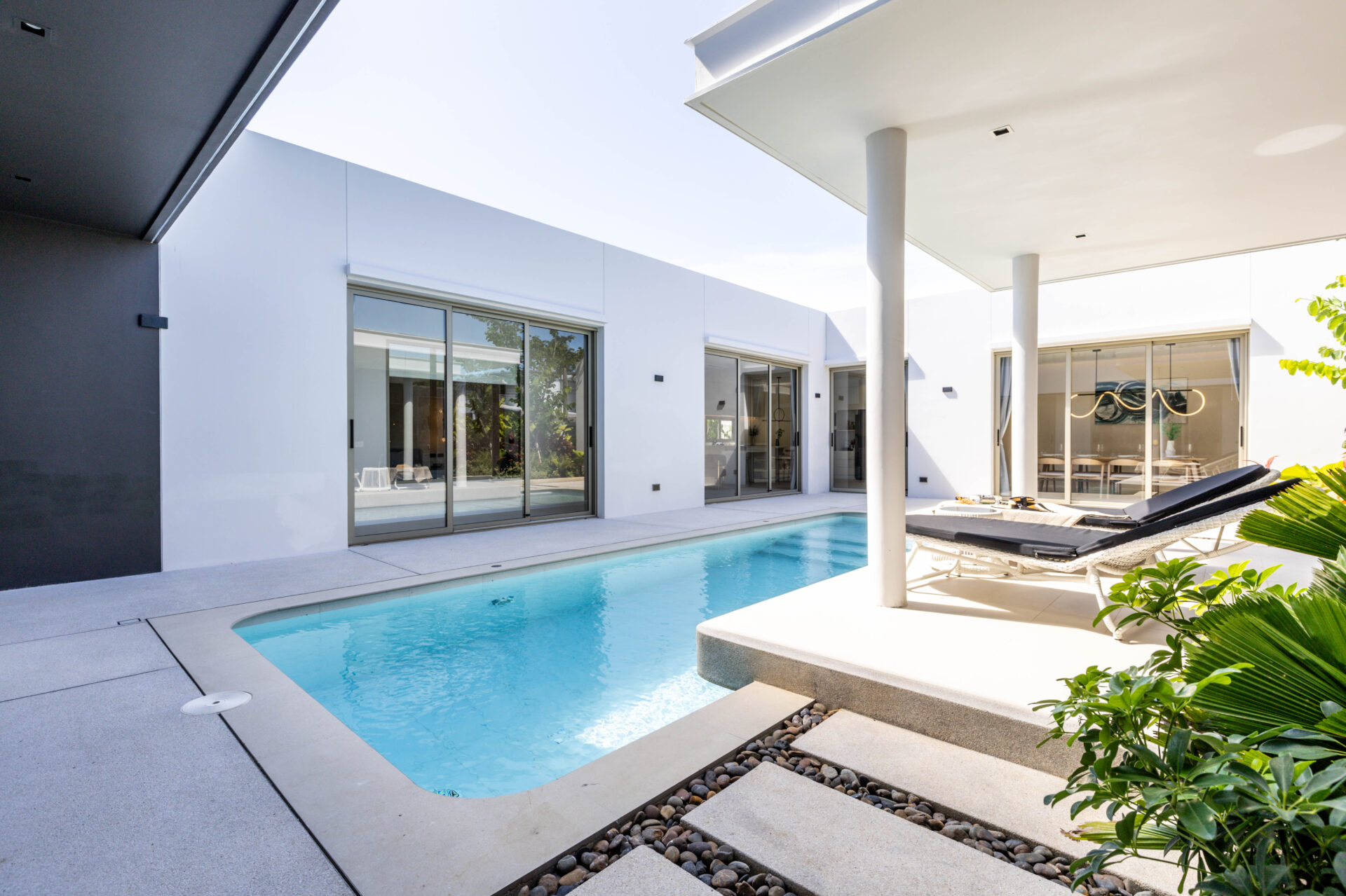 Modern white house with a rectangular pool, glass sliding doors, and sun loungers along a covered patio area.
