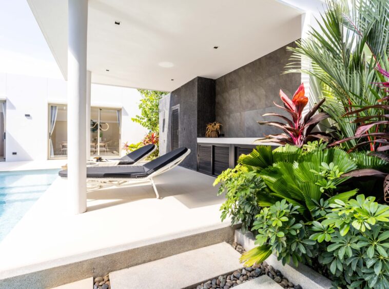 Modern poolside patio with two black lounge chairs and lush tropical plants beside a white contemporary home.