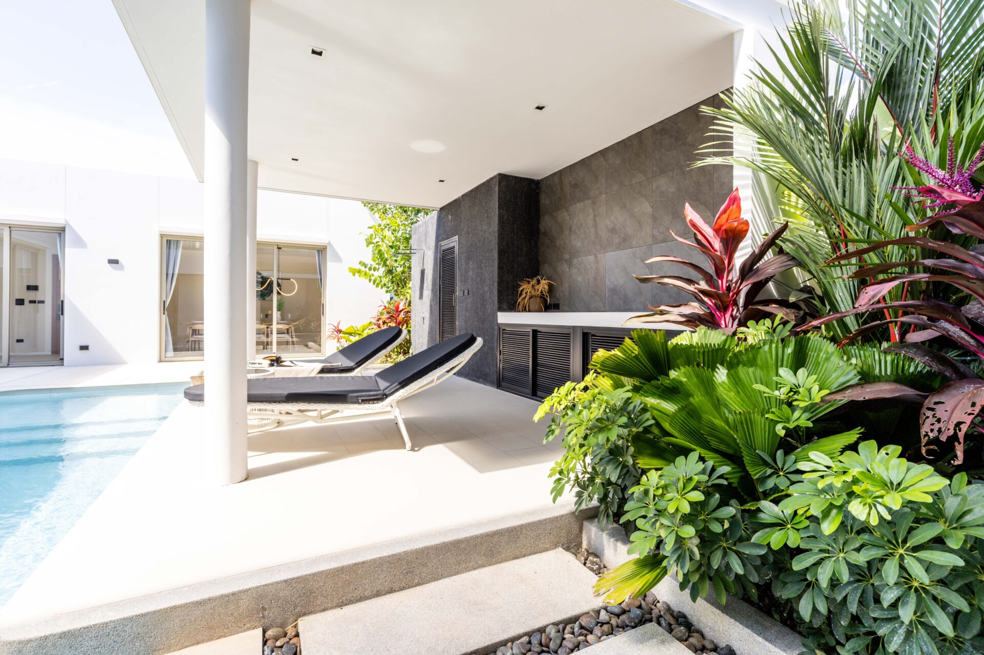 Modern poolside patio with two black lounge chairs and lush tropical plants beside a white contemporary home.