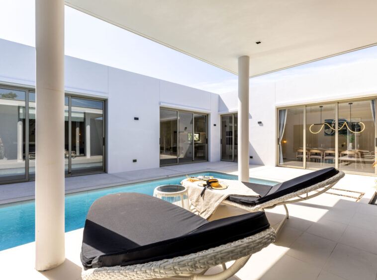 Modern courtyard with a rectangular pool, white walls, and glass sliding doors; wicker lounge beds under a shaded patio.
