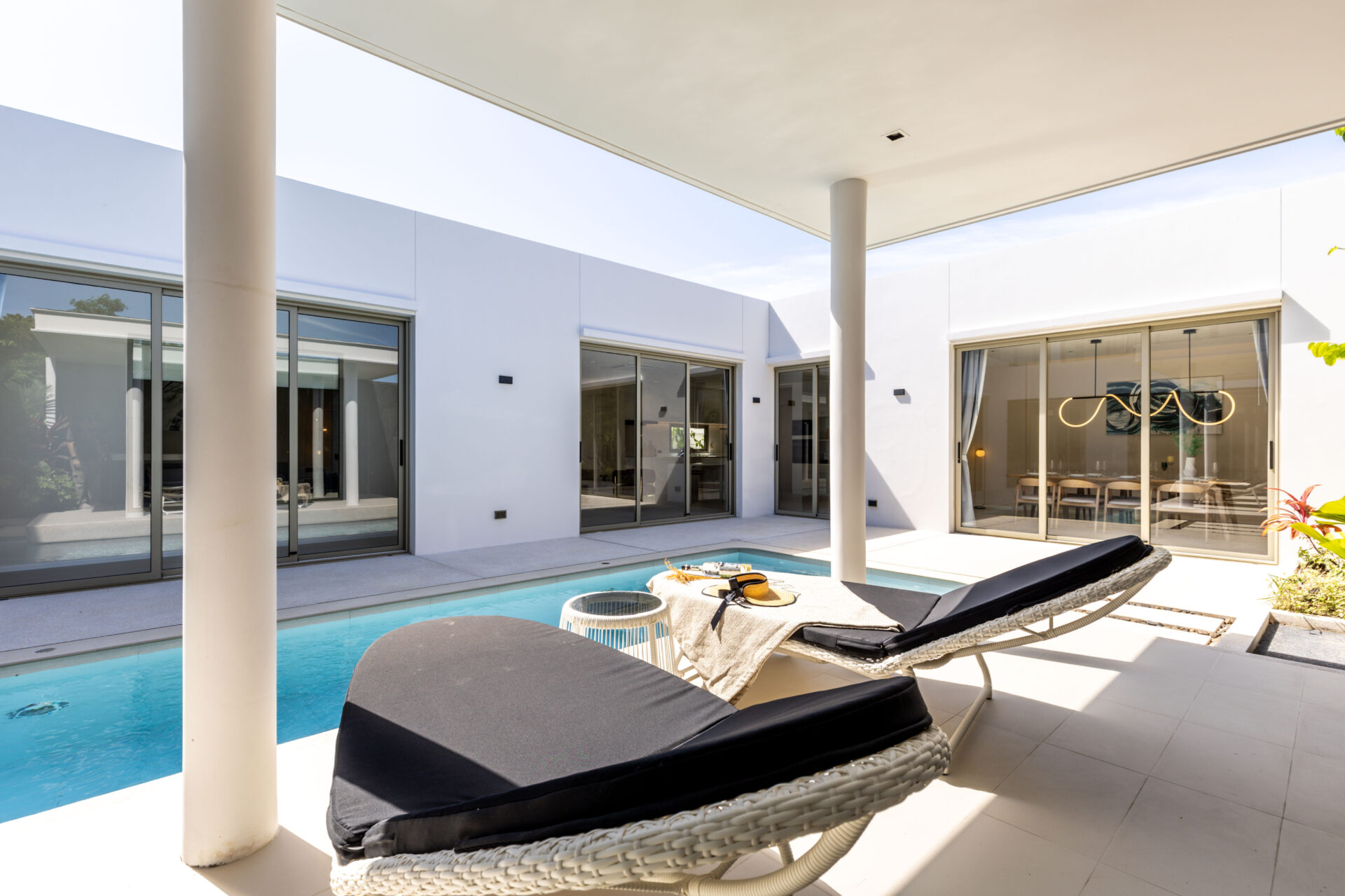 Modern courtyard with a rectangular pool, white walls, and glass sliding doors; wicker lounge beds under a shaded patio.