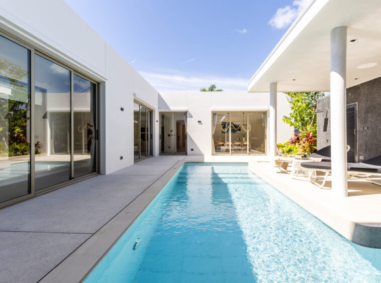 Modern white villa courtyard with glass sliding doors, a rectangular pool, and white lounge chairs.