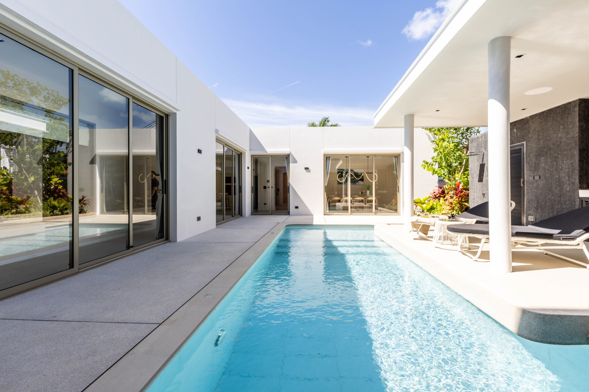 Modern white villa courtyard with glass sliding doors, a rectangular pool, and white lounge chairs.