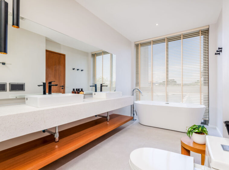 Modern bathroom with double sinks, long quartz counter, and a freestanding tub by large window with blinds