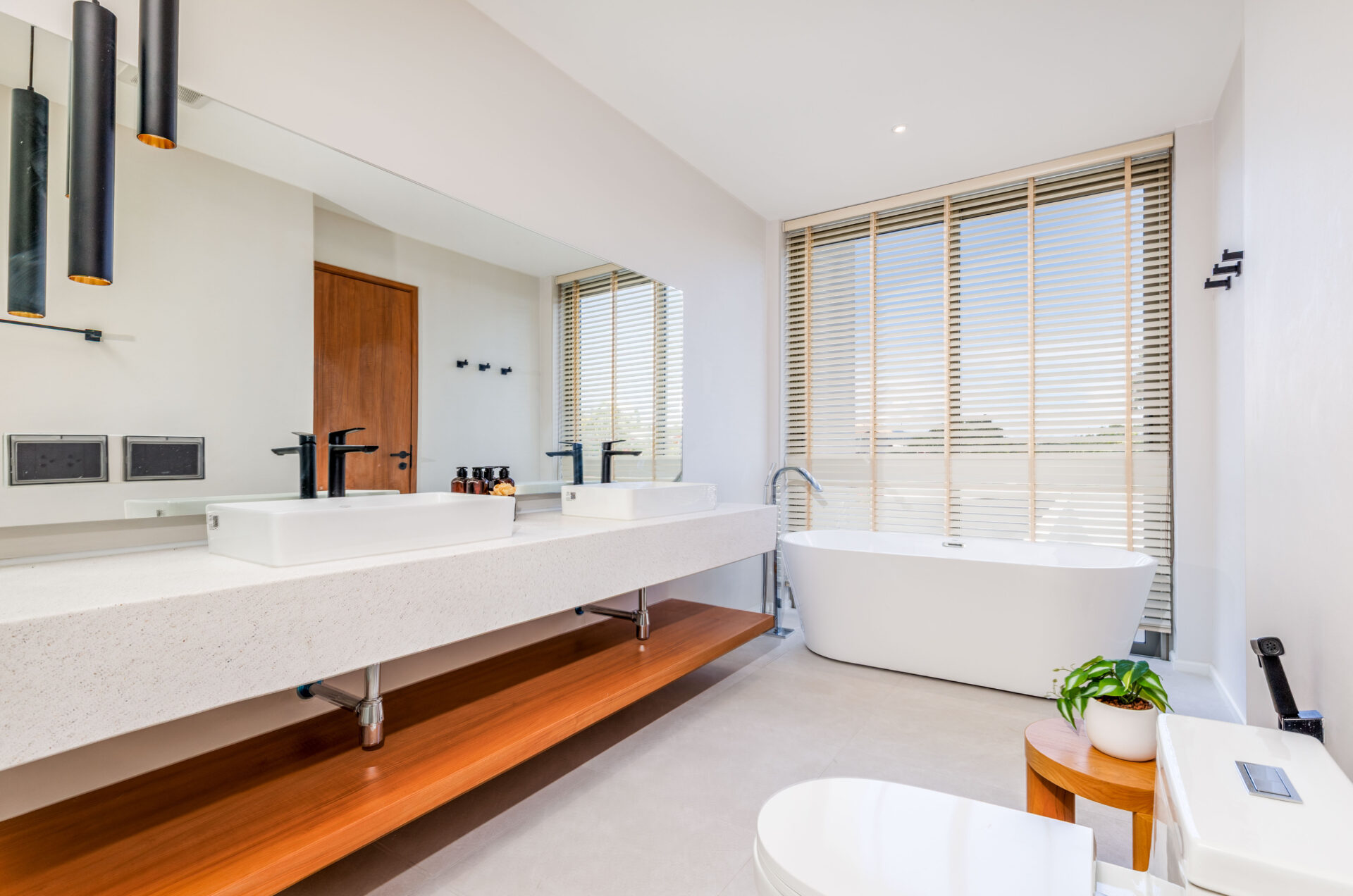 Modern bathroom with double sinks, long quartz counter, and a freestanding tub by large window with blinds