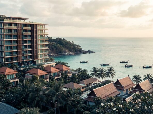 Coastal resort with a tall modern building and thatched-roof bungalows beside palm trees overlooking a calm turquoise sea. Boats anchor near the shore at sunset.
