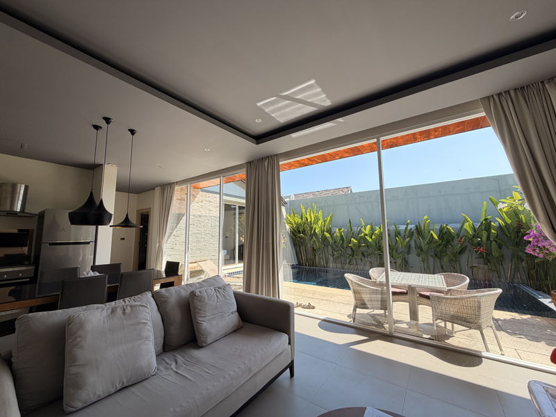 Bright living room with a grey sofa and sliding glass doors opening to a poolside patio and wicker table set.
