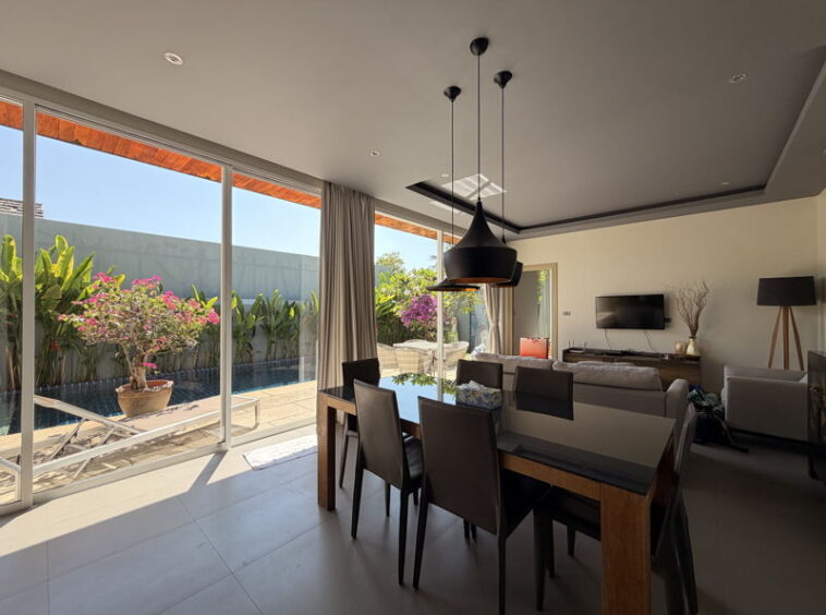 Open-plan dining and living area with glass sliding doors to a sunny outdoor patio and pool, black pendant lights overhead.