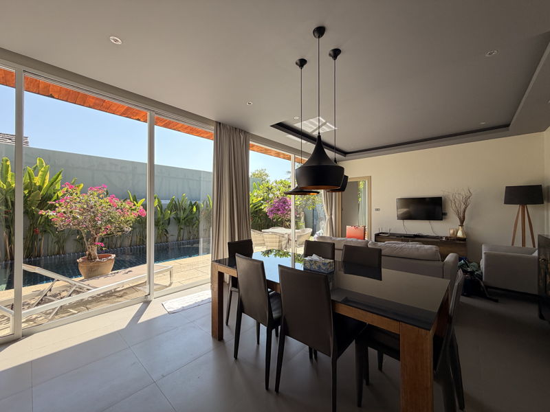 Open-plan dining and living area with glass sliding doors to a sunny outdoor patio and pool, black pendant lights overhead.
